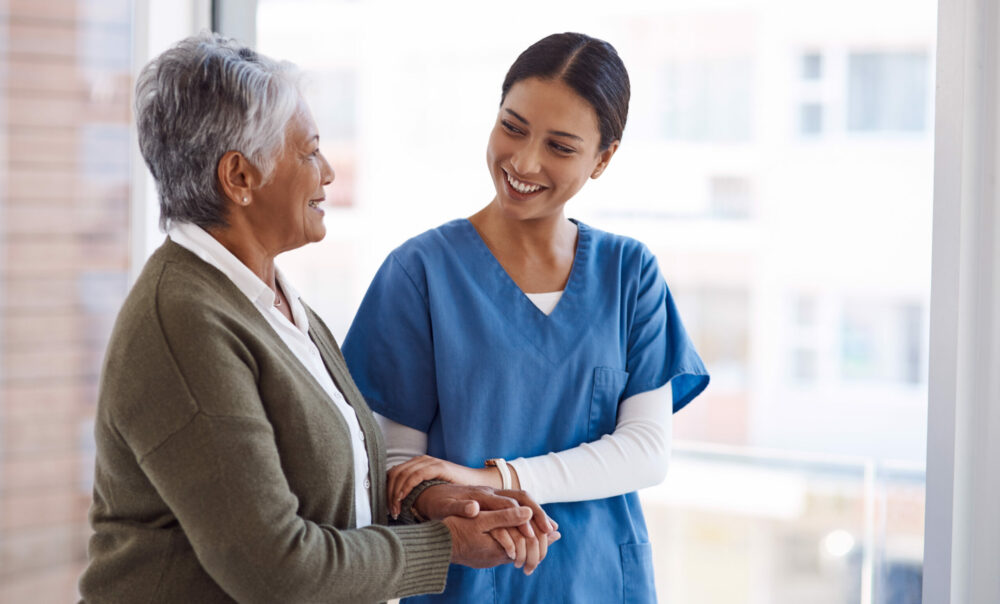 Support, caregiver with senior woman and holding hands for care indoors. Retirement, consulting and professional female nurse with elderly person smiling together for healthcare at nursing home